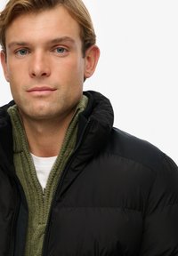 Chaqueta de plumón negra con capucha, combinada con un suéter verde texturizado debajo. La chaqueta presenta un acabado suave y acentos en los hombros de color negro.