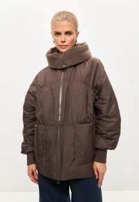Bruine pufferjack met een brede, met fleece gevoerde kraag, twee voorzakken, een ritssluiting en geplooide details langs de zijkanten.