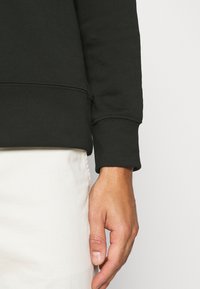 Main droite détendue le long du corps, portant un haut noir à manches longues avec poignets retroussés et un pantalon clair, sur un fond uni.