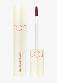 Teint Juteux Longue Durée dans un tube crème brillant avec un capuchon à vis ; il dispose d'un applicateur en forme de pied de cerf texturé et d'une teinte berry profonde.