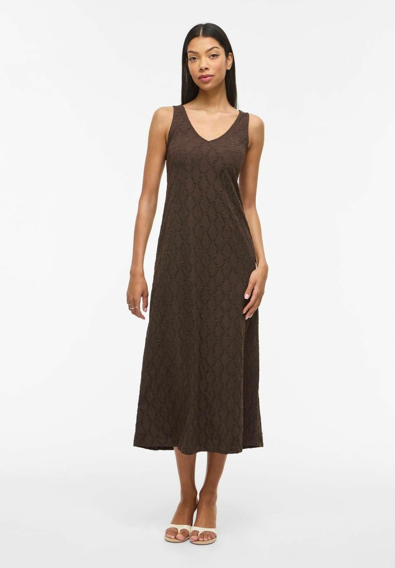 Femme portant une robe midi sans manches marron texturée avec un col en V, debout devant un fond blanc uni, chaussée de sandales beige à bout ouvert.