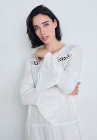 Vestido branco, leve, com mangas em sino e detalhes em renda no peito. O tecido parece suave e fluido, criando uma silhueta descontraída.