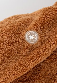 Brauner, flauschiger Stoff mit einer weichen Textur, der ein rundes Logoschild mit einem weißen Farbspritzer-Design zeigt. Das Material wirkt warm und gemütlich.