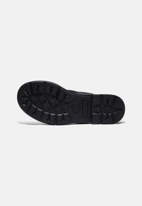 Black rubber sneaker sole with deep tread patterns, featuring a central logo and raised accents for traction. Textured and sturdy design.
