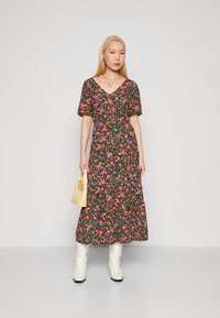 Robe florale avec un fond noir, fleurs roses et vertes, décolleté en V, manches courtes et longueur aux chevilles. Associée à des bottes blanches et un sac jaune.