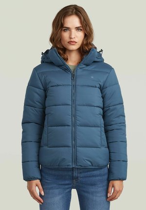 Frau mit schulterlangem braunem Haar, die eine blaue gesteppte Daunenjacke mit Kapuze und blaue Jeans vor neutralem Hintergrund trägt.