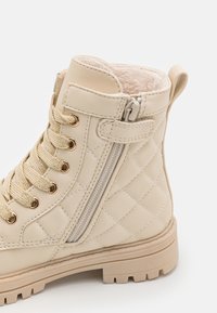 Béžová kotníková bota s prošívanou texturou, zlatými doplňky a bočním zipem. Obsahuje měkký podšívkový interiér a pevnou podrážku s vzorem.