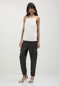 Donna che indossa un top senza maniche bianco, pantaloni cargo neri con tasche, sandali con tacco bianchi e cinturini, e grandi orecchini dorati, in piedi contro uno sfondo neutro.