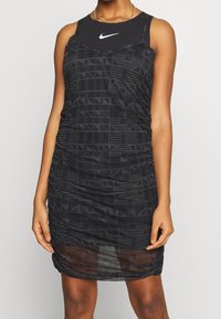 Kvinna iklädd en ärmlös svart Nike-klänning med geometriska mönster och en genomskinlig fåll, accessoriserad med ett guldhalsband.