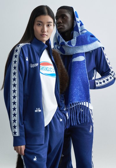 Tute blu con accenti a stelle bianche, comprensive di t-shirt con logo e sciarpa blu con frange, su uno sfondo neutro.