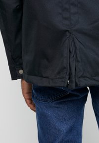Sort stofjakke med en samlet kant og sølvnapp, parret med blå denim jeans. Fokus på tekstur- og materialedetaljer.