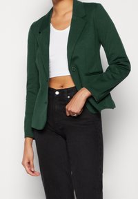 Blazer vert en tissu lisse avec une fermeture à un bouton, des revers crantés et deux poches avant, associé à un crop top blanc et un jean noir.