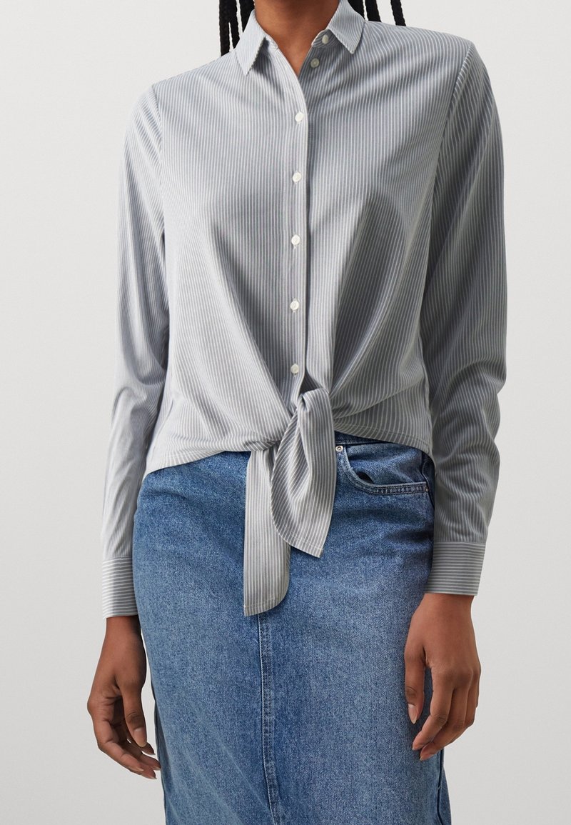 Personne portant une chemise à manches longues rayée gris clair et blanc, nouée à la taille, et une jupe en jean bleue.