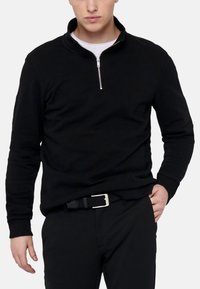 Sort halv-zip pullover lavet af et blødt materiale, med en sølvfarvet lynlås og ribkantede ærmer. Båret sammen med et sort vævet bælte og bukser.