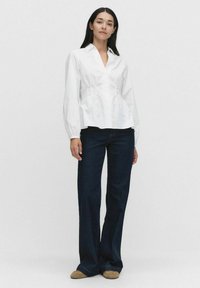 Vit långärmad skjorta med knappar och V-ringning, rynkad midja och figurnära liv, tillsammans med mörkblå utsvängda jeans.