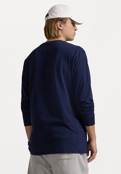 Jūrininkų ilgomis rankovėmis marškiniai iš minkštos medžiagos, su apvalia iškirpte ir aptemptomis rankovėmis, derinami su šviesiai pilkomis treniruotėmis ir balta kepure.