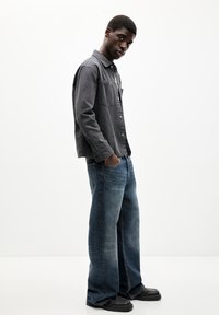 Grau button-up Hemd mit zwei Brusttaschen, kombiniert mit weit geschnittenen blauen Jeans und schwarzen Loafers, vor einem schlichten weißen Hintergrund.