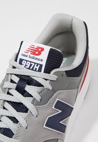Basket en suède gris et mesh avec des accents navy et des détails rouges. Caractéristiques des lacets épais et un col rembourré. Logo New Balance sur la languette.
