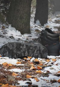 Scarponcino da trekking nero posato su un terreno forestale innevato e coperto di foglie, mostrando la suola con motivo e i tronchi degli alberi sullo sfondo.