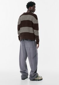 Jeune personne debout, portant un pull rayé brun et beige, un pantalon gris ample avec de grandes poches et des chaussures beiges texturées avec des lacets verts.