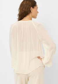 Light cream blouse in sheer fabric with long sleeves, pleated back detail, and gathered cuffs. Slightly curved hem. Paired with beige bottoms.