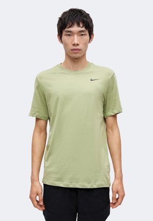 Jeune homme face à l’objectif portant un t-shirt Nike vert clair à manches courtes et un short noir sur un fond uni.