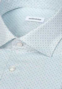 Gros plan d'une chemise habillée bleu pâle à motifs avec des boutons blancs et une étiquette "seidensticker" visible à l'intérieur du col.
