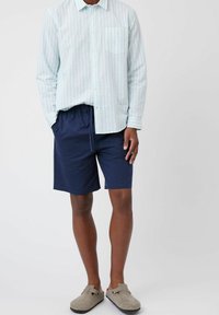 Camisa de manga larga a rayas en azul claro y blanco con un bolsillo, combinada con pantalones cortos de cintura elástica en azul marino. Zapatillas grises sin cordones.