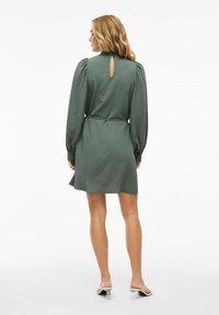 Groene, gestructureerde jurk met lange pofmouwen, een keyhole-achterkant en een getailleerde taille, die boven de knie valt, aangevuld met witte, hoge sandalen.