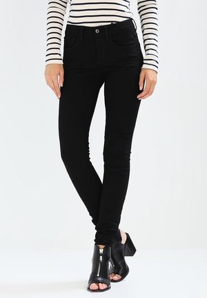 Femme portant un jean skinny noir, un haut à manches longues rayé et des bottines noires à bout ouvert avec fermeture éclair devant, debout jambes croisées.