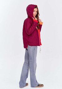 Kobieta w bordowej pikowanej kurtce z kapturem, pomarańczowym swetrze, jasnoniebieskich dżinsach i butach sportowych w panterkę, stojąca w profilu.