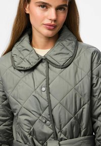 Chaqueta verde acolchada con un gran cuello, cierres de botones y una cintura con cinturón. Presenta un patrón de diamantes y una textura de tela suave y brillante.