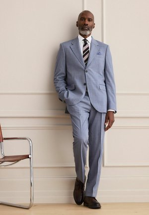 Mann mit grauem Bart in hellblauem kariertem Anzug, gestreifter Krawatte und braunen Schuhen lehnt an einer cremefarbenen Wand neben einem Metall- und Lederstuhl.