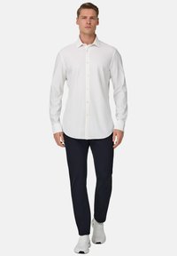 Weißes Hemd mit Knopfleiste, lange Ärmel, glatter Stoff, gerader Saum, kombiniert mit dunkler Hose und weißen Turnschuhen. Einfaches Design mit dezenten Akzenten.