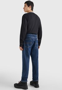 Camisa negra de manga larga, pantalones de mezclilla azul con dos bolsillos traseros y zapatos negros gruesos. Los pantalones de mezclilla tienen un acabado texturizado y costuras visibles.