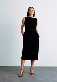 Svart ärmlös klänning med rak skärning, texturerat tyg, sidfickor och knytaljett i midjan. Bärs med svarta högklackade sandaler.