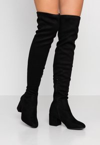Botas negras sobre la rodilla, hechas de ante, con un tacón cuadrado, punta afilada y detalles de cremallera lateral. Textura suave y ajuste elástico.