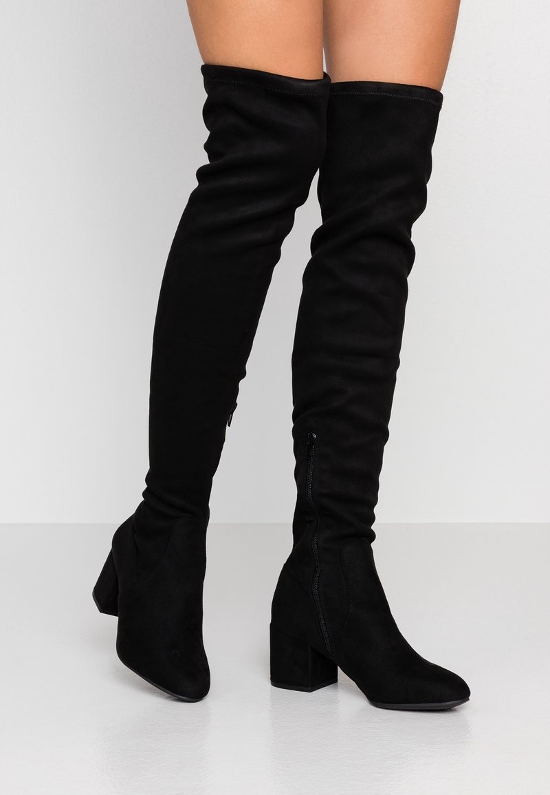 Botas negras sobre la rodilla, hechas de ante, con un tacón cuadrado, punta afilada y detalles de cremallera lateral. Textura suave y ajuste elástico.