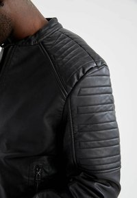 Veste en cuir noir avec accents matelassés aux épaules, fermeture éclair et texture lisse. Présente une coupe ajustée et un style minimaliste.