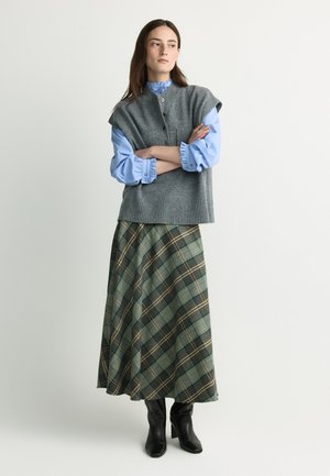 Woman with long brown hair wears a gray sleeveless sweater over a blue ruffled blouse, a green plaid skirt, and black heeled boots standing with arms crossed.