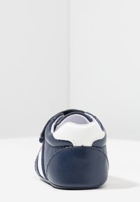 Námořnické kožené boty s bílou patní klapkou, suchým zipem a červenými a bílými bočními pruhy; hladká textura a zaoblený design.