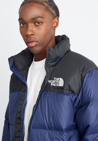 Puffjakke i marineblå og sort med høj krave, der har lynlås lukning og "The North Face" logo på brystet. Glat, skinnende overflade.