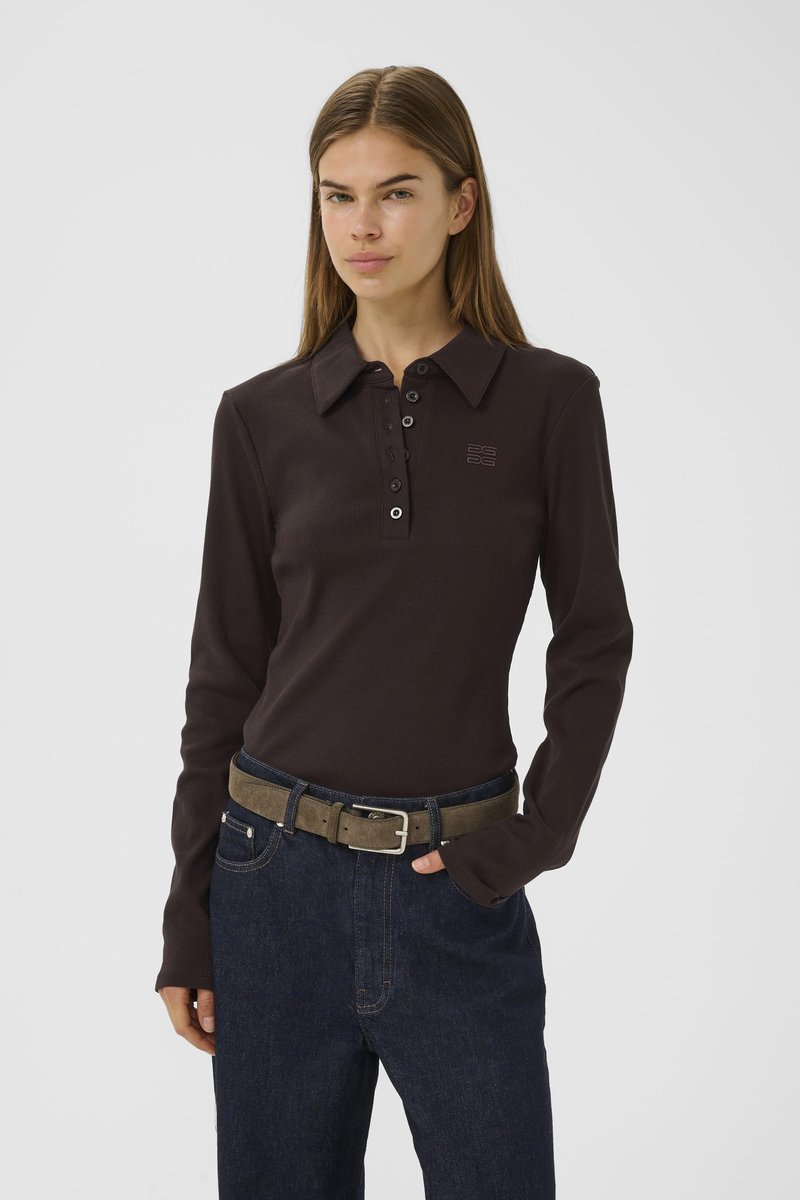 Woman wearing a brown long-sleeve polo shirt with buttons and dark blue jeans, standing with one hand in pocket against a plain background.