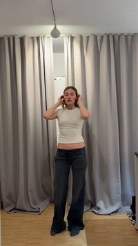 Mladá žena si upravuje vlasy, stojí bosá na dřevěné podlaze před zavřenými béžovými závěsy s osvětlením na stropě nad ní.