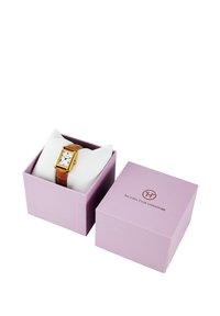 Montre rectangulaire dorée avec cadran blanc et marqueurs noirs, montée sur un bracelet en cuir marron. Emballée dans une boîte rose avec logo.