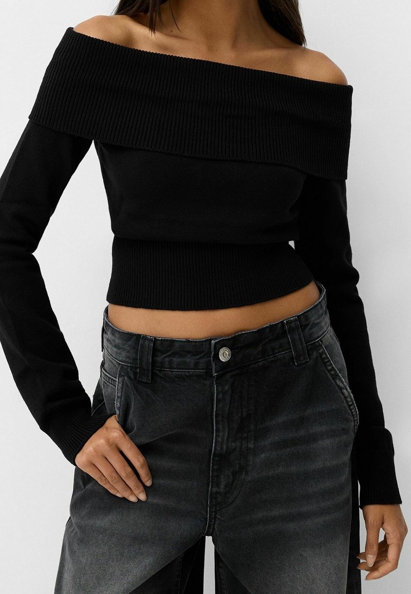 Femme portant un pull noir côtelé à épaules dénudées et un jean noir délavé taille haute, main posée sur la hanche, sur fond uni.