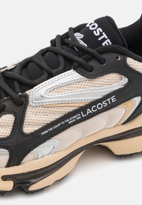Lacoste sportcipők hálós felsőrésszel, fekete, bézs és ezüst díszítésekkel. Fekete cipőfűzők, párnázott talp és márkázott heveder részlet.