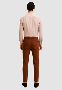 Camicia a maniche lunghe rosa chiaro con polsini abbottonati, abbinata a pantaloni tailored marroni, vista da dietro. Tessuto liscio e vestibilità slim.
