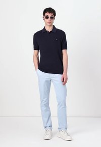 Polo-shirt bleu marine à côtes avec manches courtes ; chinos bleu clair ; baskets blanches ; lunettes de soleil rondes ; modèle posant contre un fond uni.