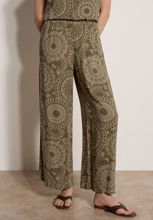 Person trägt olivgrüne, weite Hose mit hellen, geometrischen Mandala-Mustern und braune, gekreuzte Sandalen, steht auf beigem Boden.